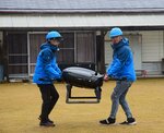 約１３キロを飛行して着陸したドローンを運ぶ関係者ら＝６日、琴浦町古長の古布庄地区公民館