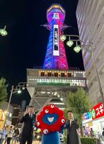 　万博のイメージカラーにライトアップされた通天閣の前で、公式キャラクターのミャクミャクと記念撮影する「通天閣観光」の西上雅章会長（右）ら＝２４日夜、大阪市浪速区