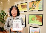 絵本「ぼく　どうなるの？」を持つ昌子さん。壁面の絵は昌子さんが手がけた挿絵の原画