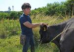 　牧草の生い茂る牧場で牛の世話をする小嶺博泉。後方の山上に見えるのは自衛隊が設置したアンテナ群＝２０２４年９月、沖縄県与那国町