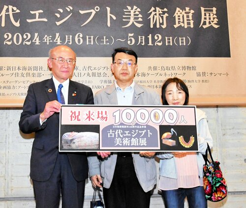 来場者１万人目の記念プレートを手にする靖弘さん（中央）と郁代さん（右）＝１９日、鳥取市の県立博物館